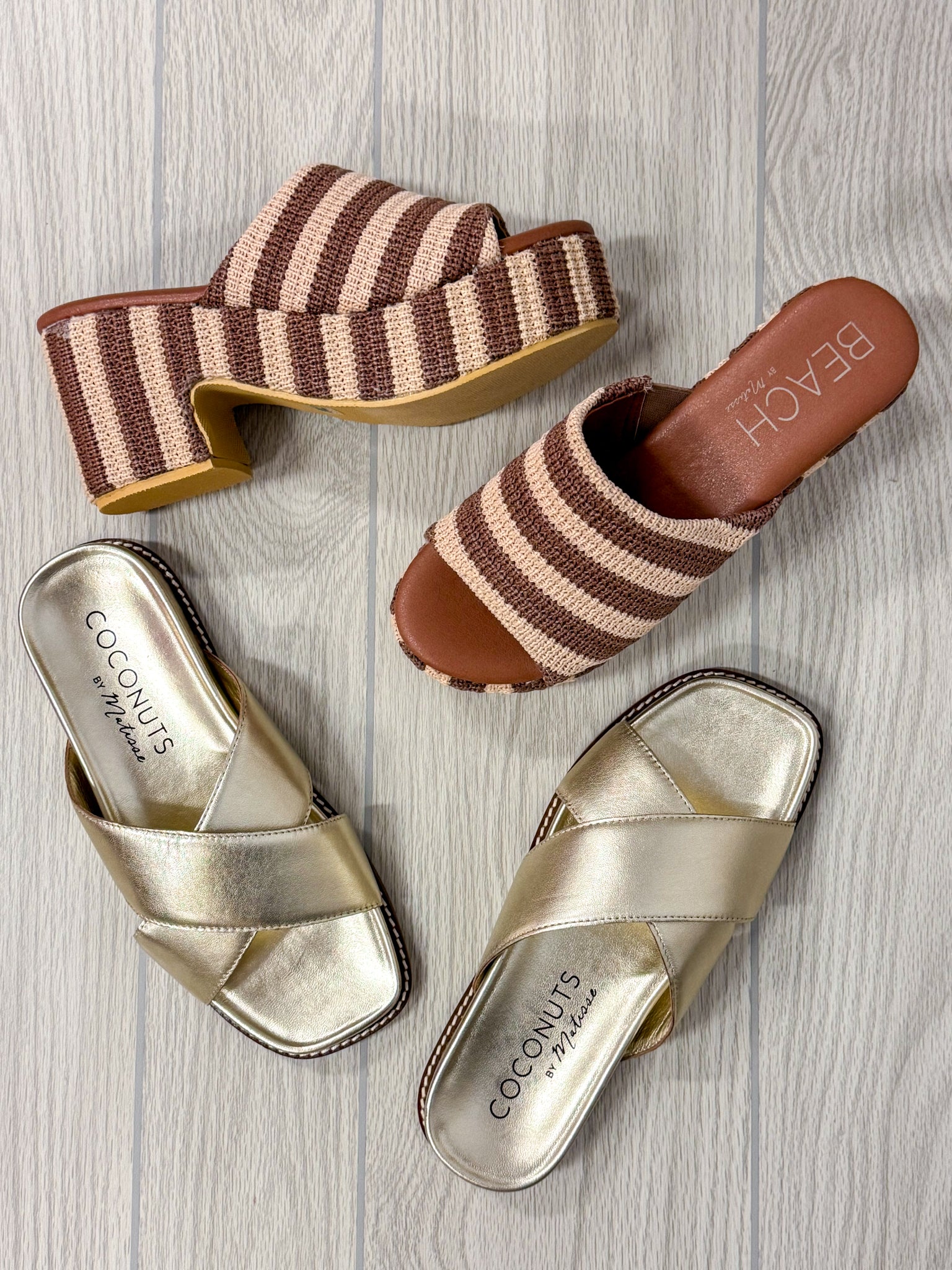 Four women's slide sandals arranged on a light gray wood floor: two brown-and-beige striped platform slides and two metallic gold crisscross slide sandals. Brand names 'COCONUTS by Nicole' and 'BEACH' are visible on the insoles.