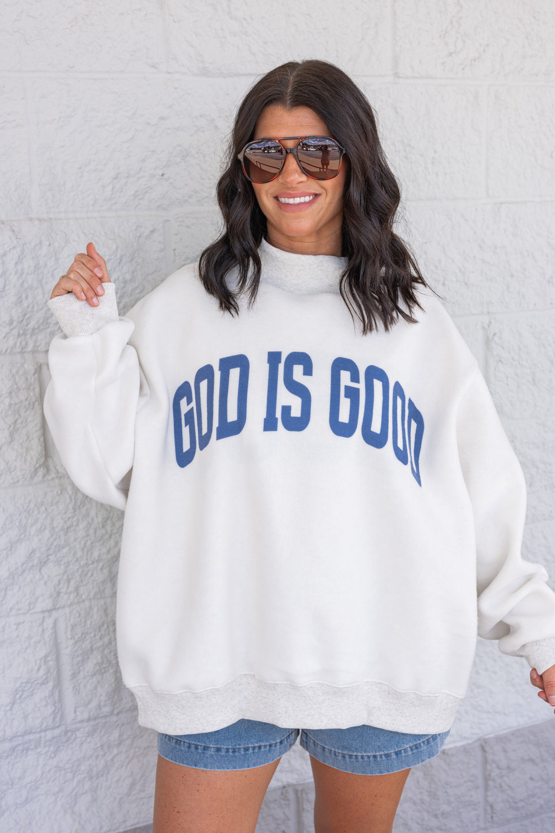 Woman wearing a white sweatshirt with 'GOD IS GOOD' text against a light gray wall.