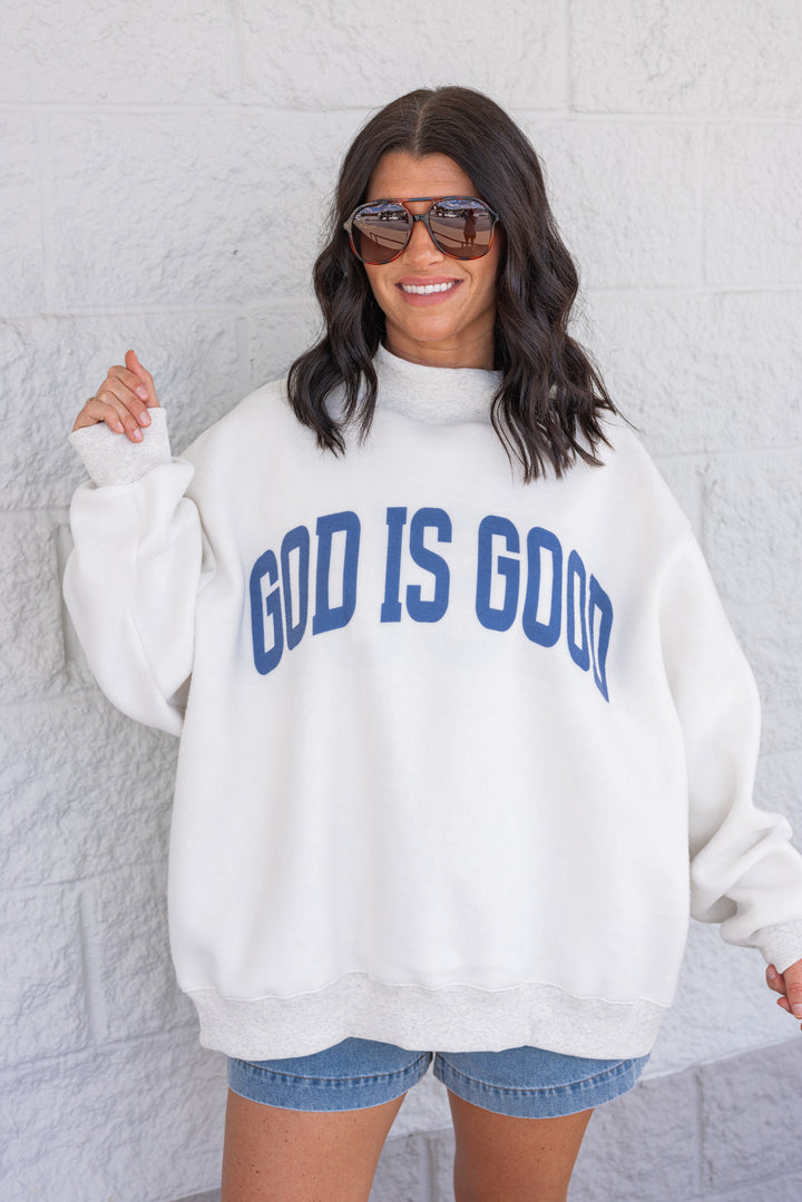 Woman wearing a white sweatshirt with 'GOD IS GOOD' text against a light gray wall.